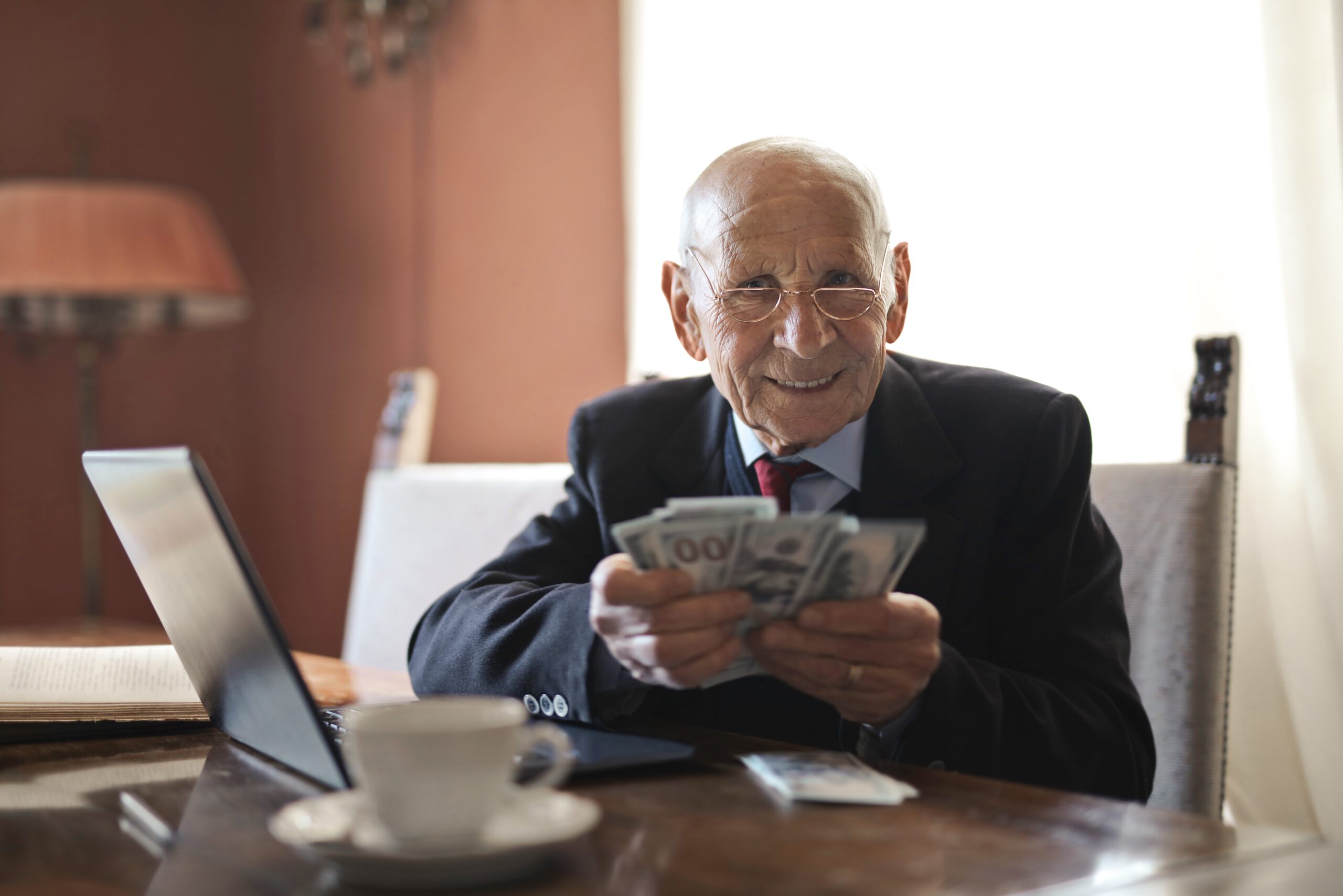 Starszy mężczyzna w okularach, siedzący przed biurkiem i liczący trzymane w dłoni banknoty z inwestycji
